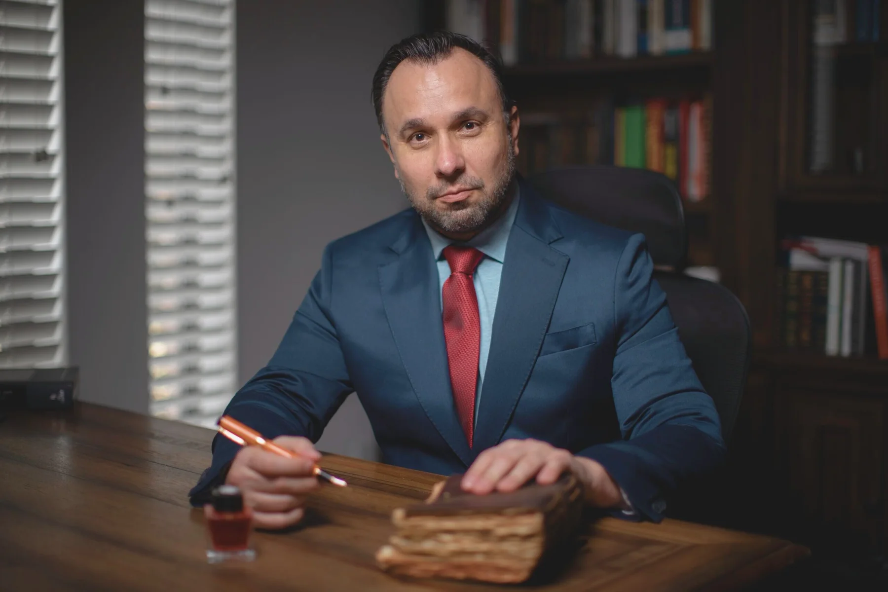 Dr. Daniel Toledo à mesa, de terno azul, assinando com caneta-tinteiro diante de uma estante